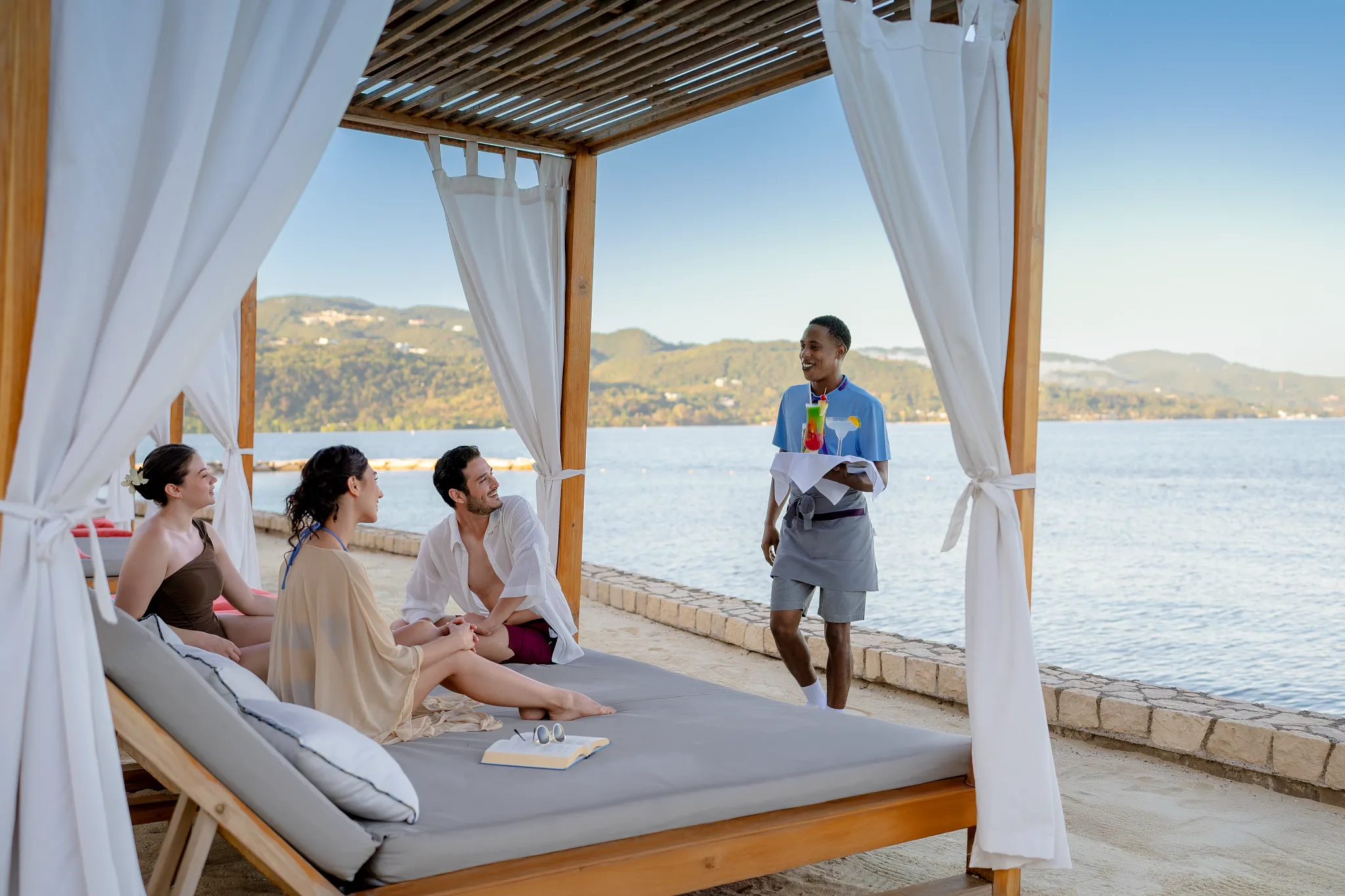 Guests relaxing on a beachfront Bali bed with cocktails at Secrets Wild Orchid Montego Bay in Jamaica.